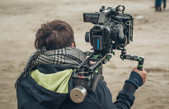 A camera operator on location with a shoulder-mounted rig, facing away from the camera. They are looking at the viewfinder and focusing on the subject