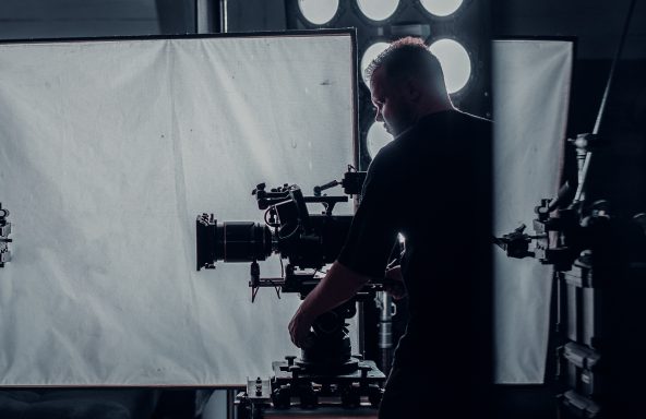 Director of photography with a camera on a tripod in front of him. The set is dimly lit with some mechanical equipment as the focus.