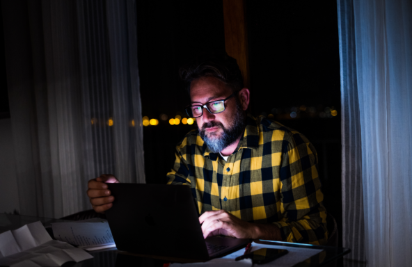 A behind-the-scenes industry worker is sat working on a laptop late at night. The light from the screen is shining on to their face.