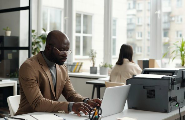 A male office worker is sat at a desk, working on a laptop in a bright modern office.