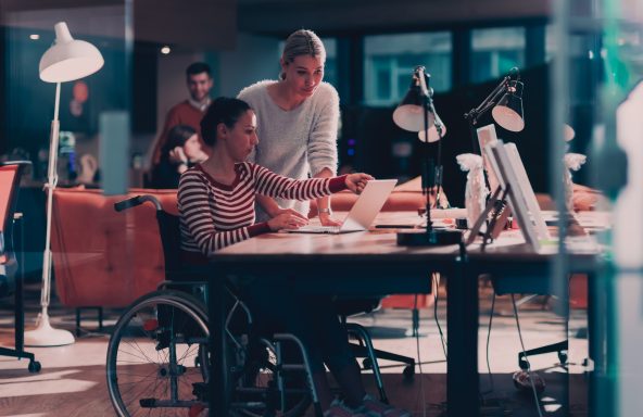 Two women at a desk, one is in a wheelchair and the other standing by their side. They are both looking at the laptop screen.