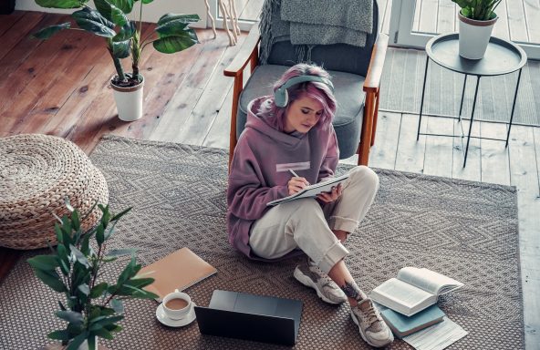 A woman sat on the floor in a living room wearing headphones and writing in a notebook. In front of her is a laptop and a pile of books.