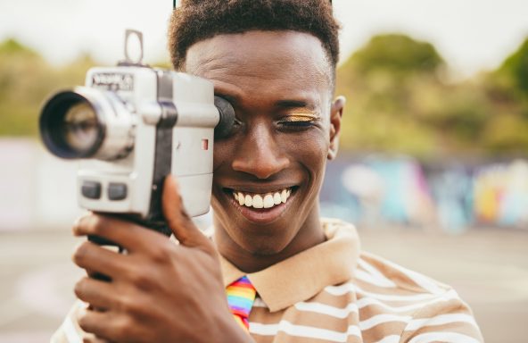 A young filmmaker wearing a brown and white striped polo shirt and a rainbow colour tie is holding a Super 8 camera viewfinder up to their eye. They have a wide smile on their face.