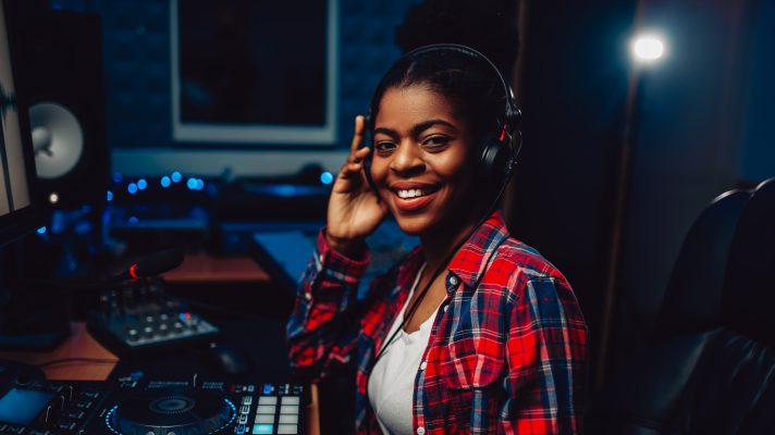 A female audio editor is sitting at a sound desk wearing headphones, she is smiling at the camera.