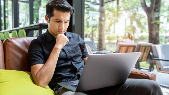 A young man is sat on a sofa. On his lap he has a laptop that he is attentively watching.