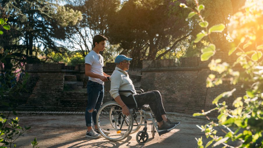 A man pushing an older man in a wheelchair through a park.