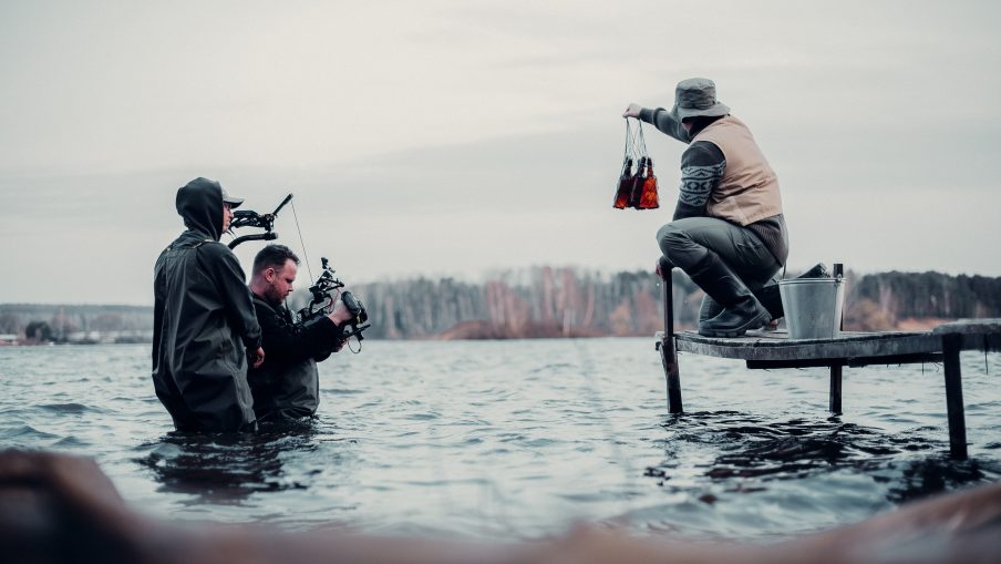 A camera operator and their assistant is standing waist-deep in lake water whilst filming a fisherman that is crouching down on an old pontoon. In the distance is a woodland area on the other side of the lake.