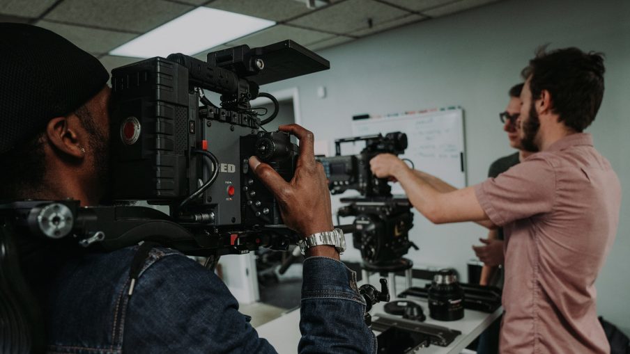 Close up of a camera operator holding a RED camera. The camera is pointing at two other crew members who are prepping an additional camera.