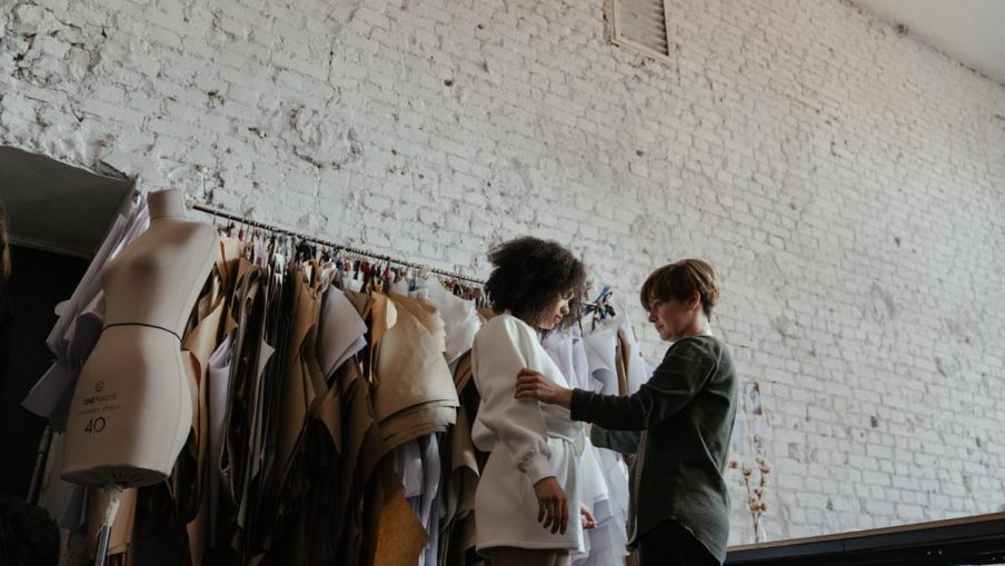 A female costume designer is in a bare, white-brick studio with a rack of clothes hanging behind them. A female model is being fitted with a white over-sized jumper and skirt.