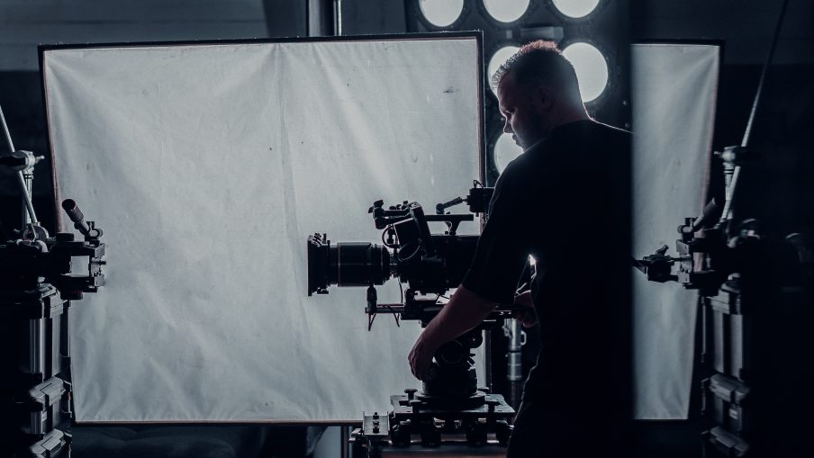 Director of photography with a camera on a tripod in front of him. The set is dimly lit with some mechanical equipment as the focus.