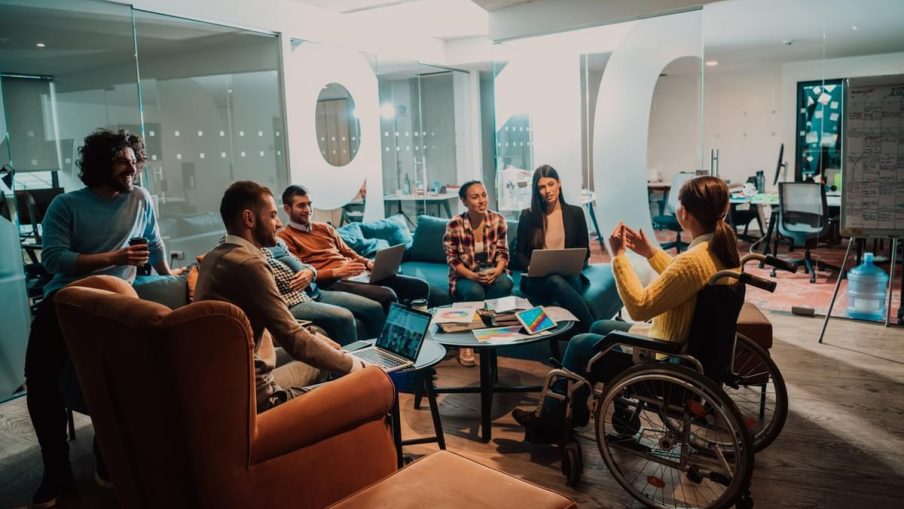 A group of people are sat in an modern and bright office meeting space. One woman in a wheelchair is sat at the front, addressing the others.