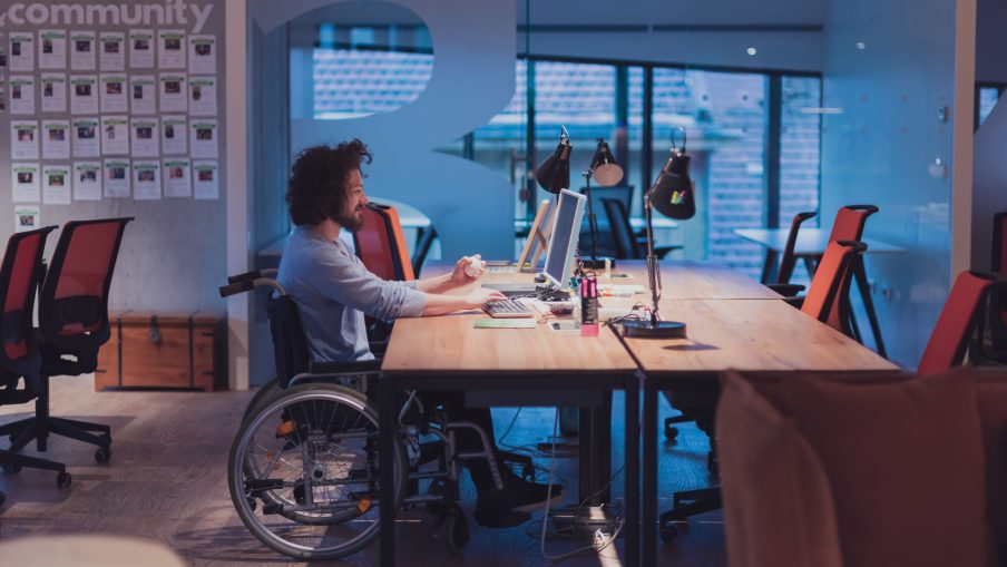 A man working at a desk in wheelchair and using a desktop computer in an open space modern office.