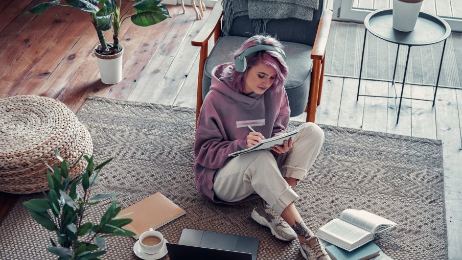 A woman sat on the floor in a living room wearing headphones and writing in a notebook. In front of her is a laptop and a pile of books.