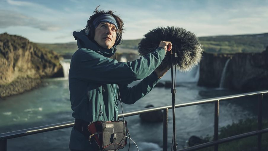A male boom operator is setting up their equipment. They are wearing cold weather gear whilst standing on an open bridge with a river below.