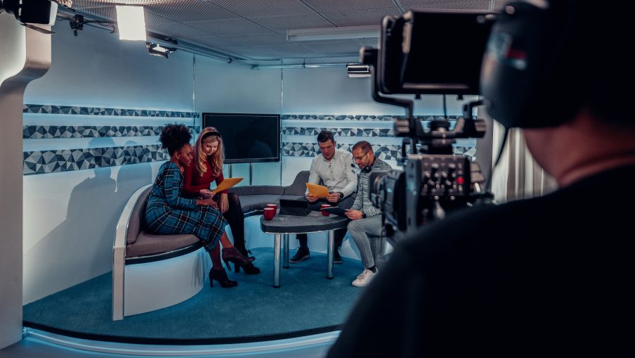 Over the shoulder view of a TV show in the process of being filmed in a studio. The presenters are sitting on the studio sofa while looking at notes and talking to the producer, while the camera man is preparing to start filming again.