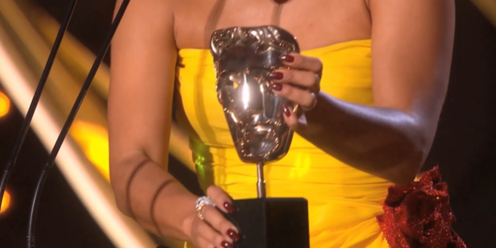 Close up of a BAFTA award being held by a woman wearing a bright yellow dress during an acceptance speech.